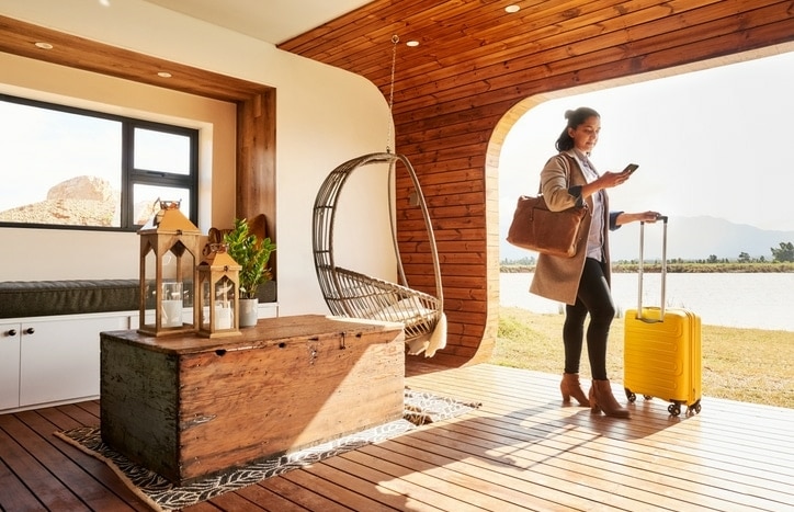 Young woman with a suitcase using her mobile phone while standing at lounge of her vacation home during a trip